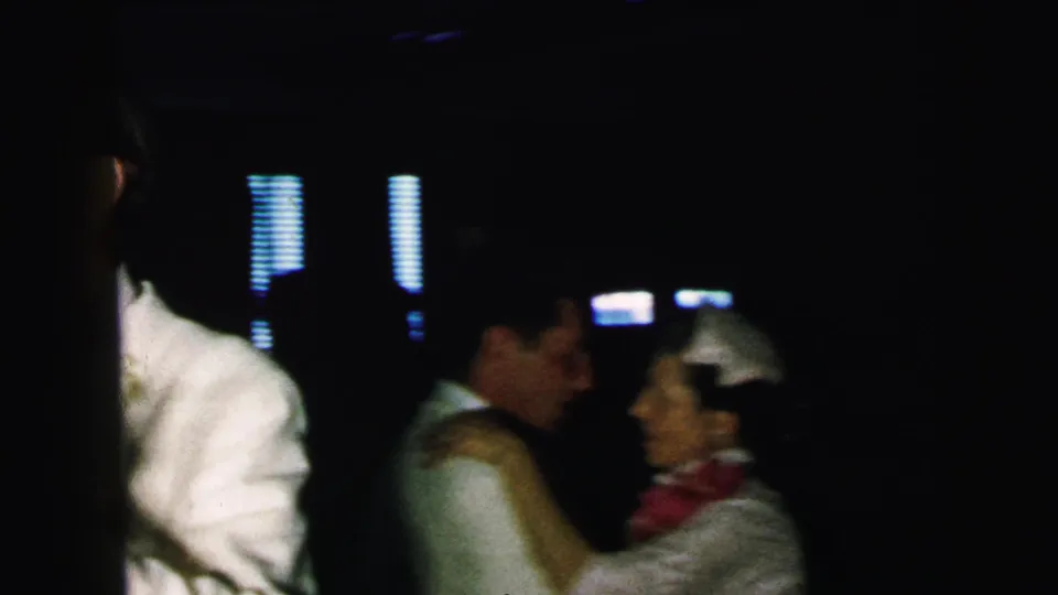 Dim 1964 Camden indoor dance scene with formal guests, a white dinner jacket, and a woman in a head covering or light accessory near the right.