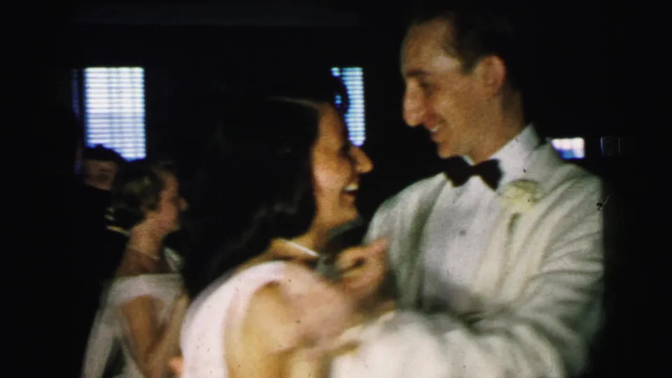 1964 Camden home-movie frame of a smiling man in a white dinner jacket dancing with a woman in a pale dress indoors.