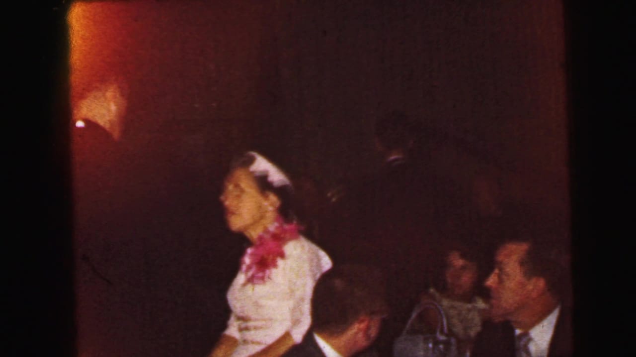A woman wearing a light outfit and pink floral accessory appears near seated guests in dim, reddish 1964 Camden wedding reception footage.