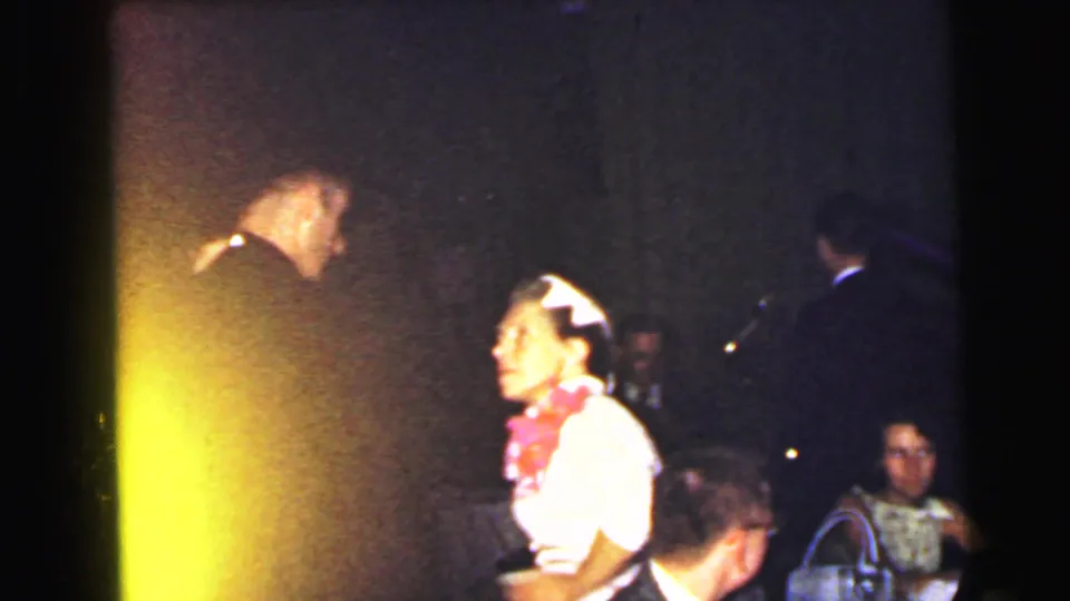 In the 1964 Camden wedding reception, a man in a dark suit faces a woman with a pink floral accessory while guests sit behind them.