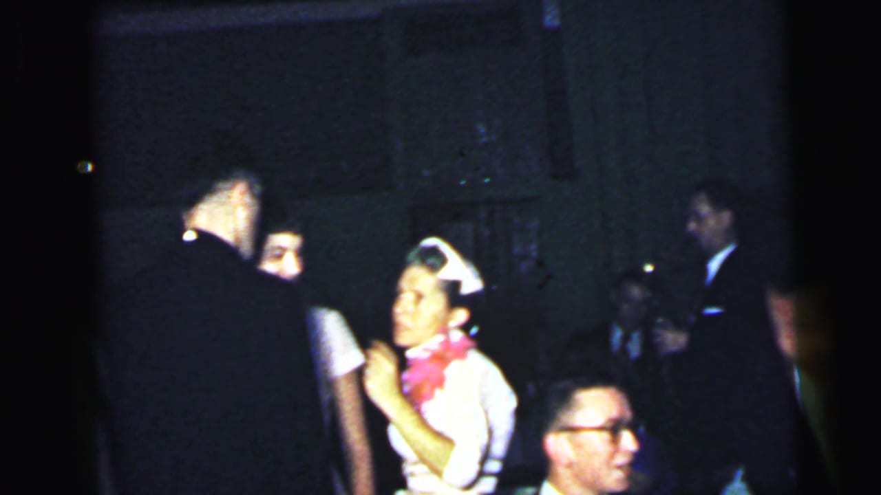 Wedding guests in dark suits and white caps gather in a dim indoor room.