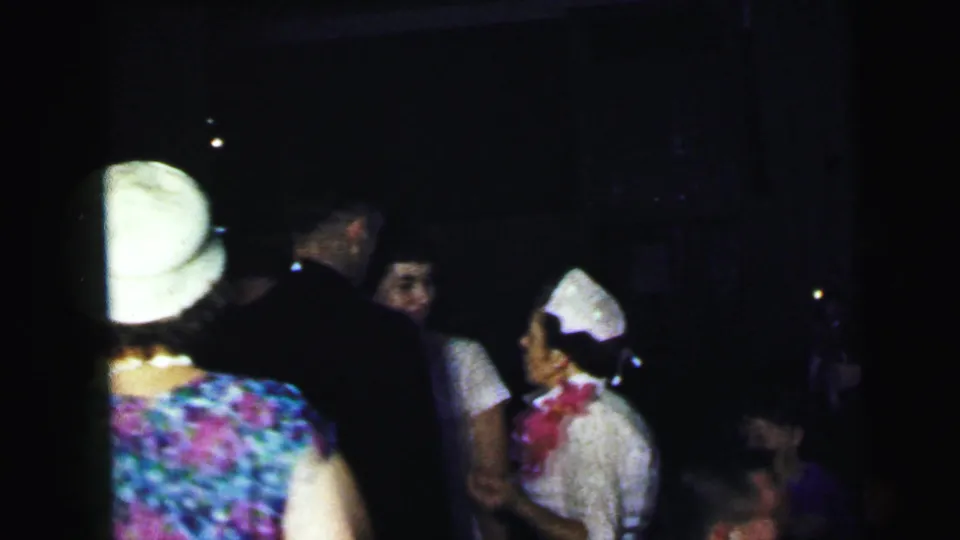 Guests at the Camden wedding reception stand in a dark room, with a floral dress, white hat, dark jackets, and a woman in a white cap visible.