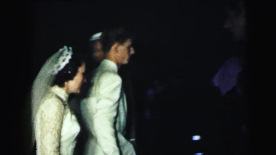 At the Camden wedding, the bride in a lace dress and veil walks beside the groom in a light-colored suit through the dim interior.