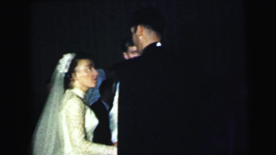 The bride in a veil and lace gown is flash-lit while facing a dark-suited man during the 1964 Camden wedding celebration.
