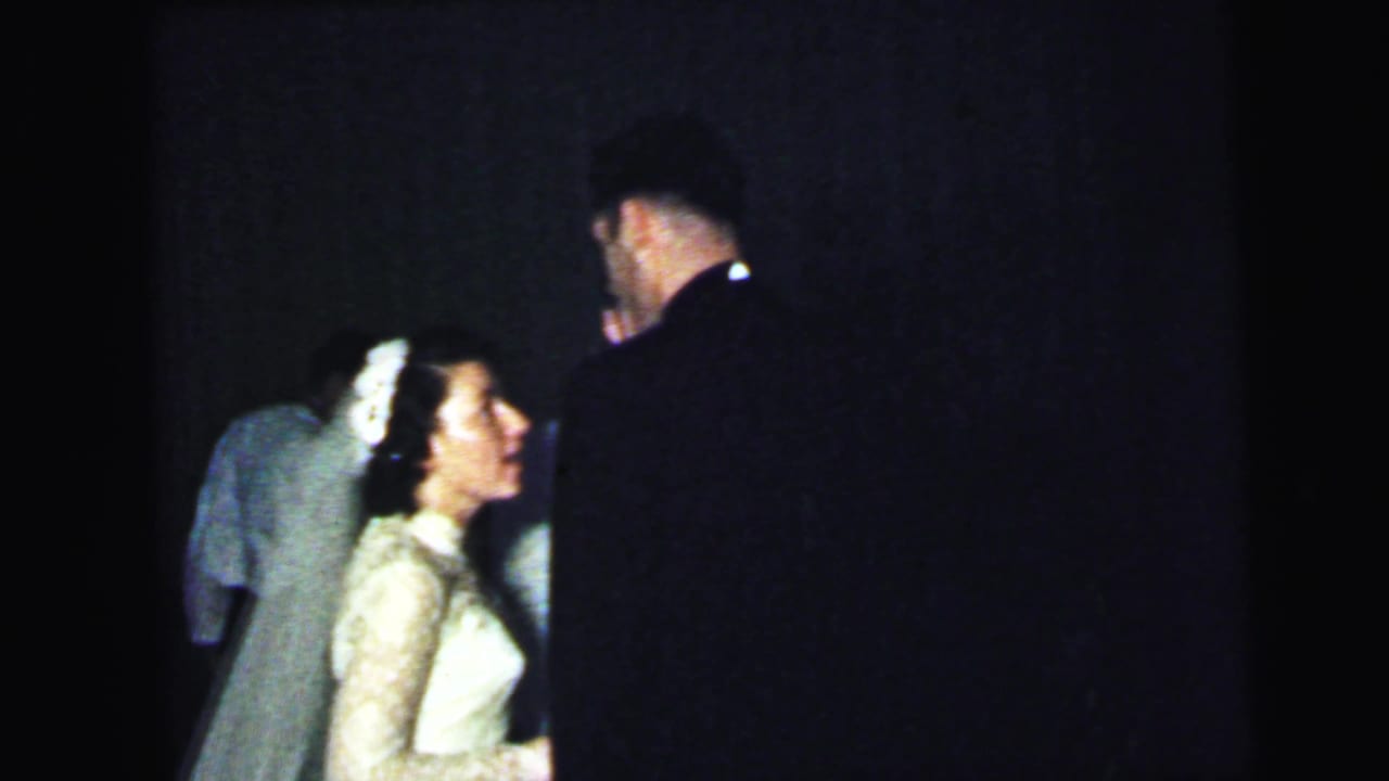 Bride and dark-suited guest beside other attendees in a dim reception room.