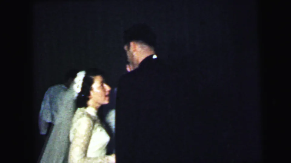 In 1964 Camden wedding footage, the bride in a white veil stands near a dark-suited guest and other attendees in low indoor light.