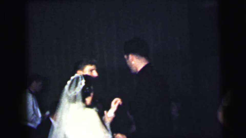 The bride's veil and gown are seen from behind as she talks with a guest in the dim 1964 Camden wedding reception footage.