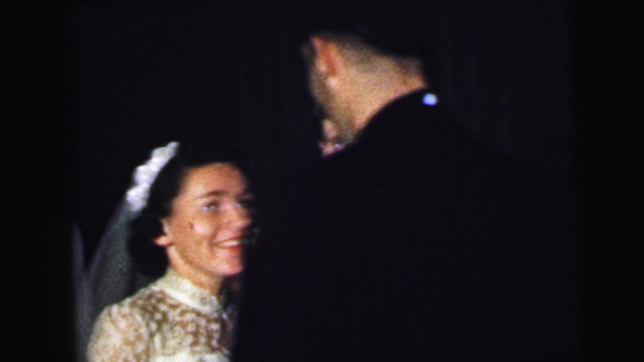 At a 1964 Camden, New Jersey wedding reception, the bride smiles in a lace dress and veil while facing a man in dark formalwear.