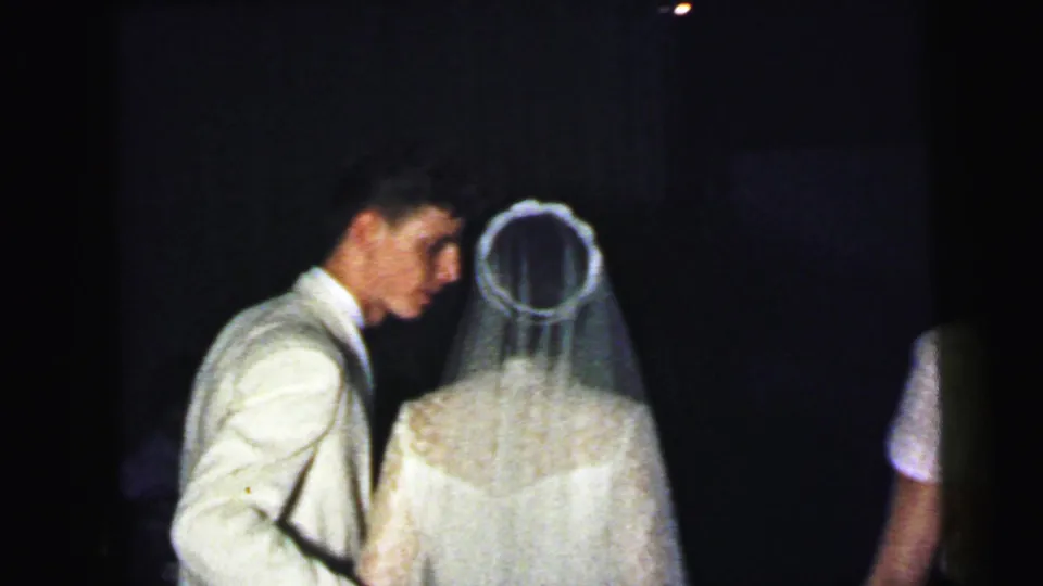 The groom in a light jacket faces the bride, whose veil and lace back are visible, during the 1964 Camden wedding reception.