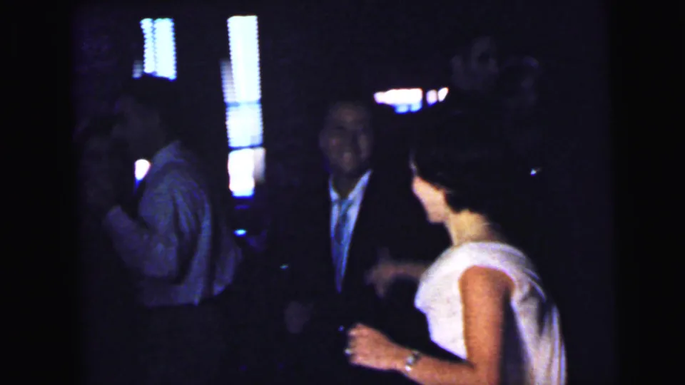 Guests in formal clothing mingle in a dim Camden reception room while the bride or a female guest in a light dress stands at right.