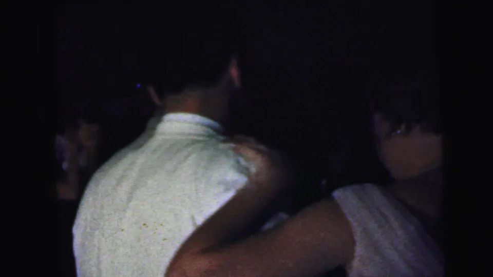 A woman places an arm around a man in a light shirt while other guests are partly visible in the dim 1964 Camden party footage.