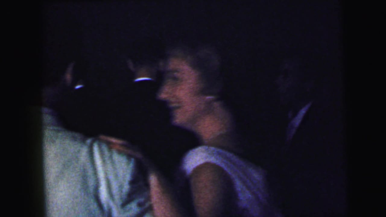 Dim party scene with adults close together, including a woman in a sleeveless light dress.