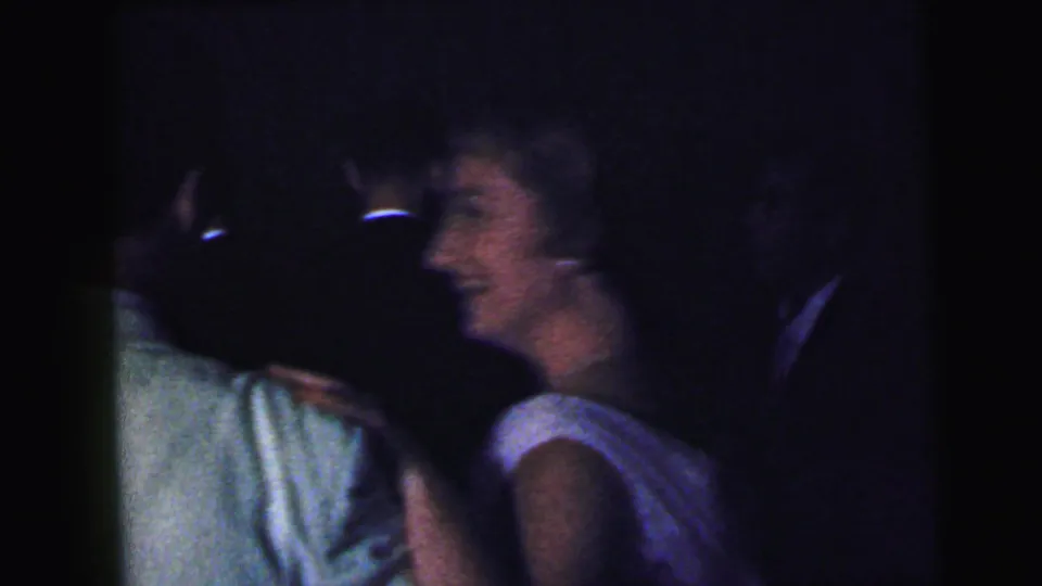1964 Camden home-movie frame showing adults dancing or socializing closely in a dark interior, with soft focus and heavy grain.