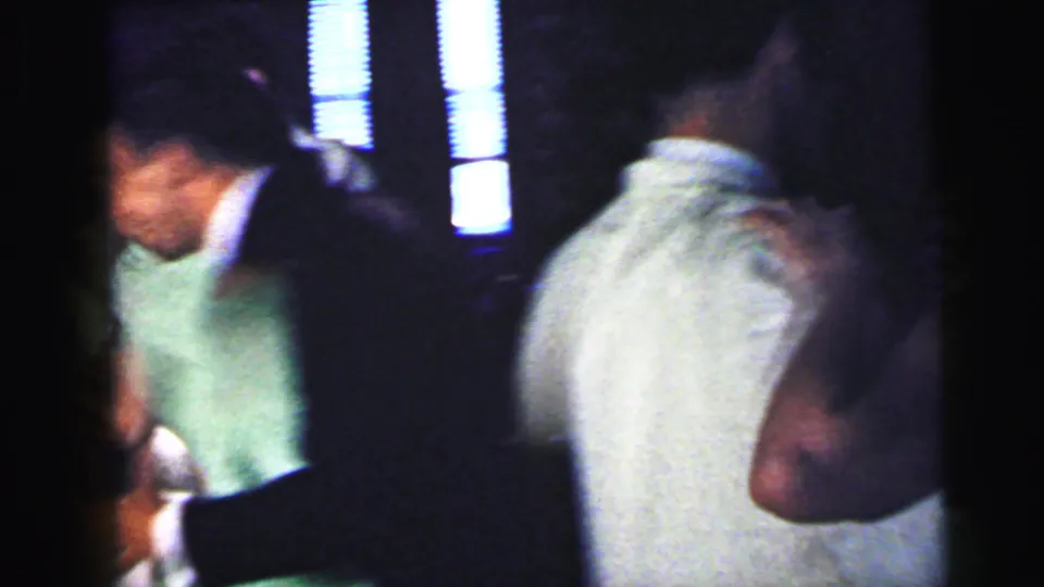 1964 Camden party frame showing several adults in close quarters, including light shirts and bright vertical window light in the background.