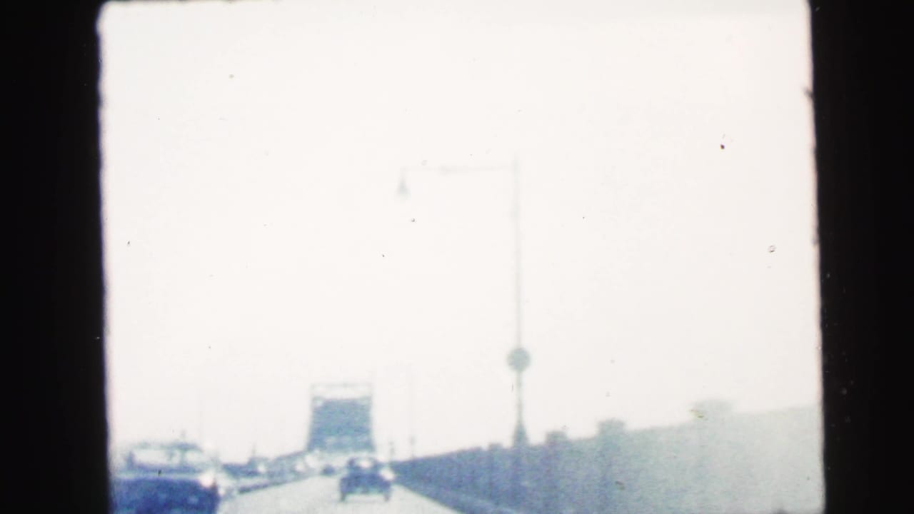 1964 home-movie frame from Camden, New Jersey showing a bright highway view from inside a moving car, with traffic and roadside structures ahead.