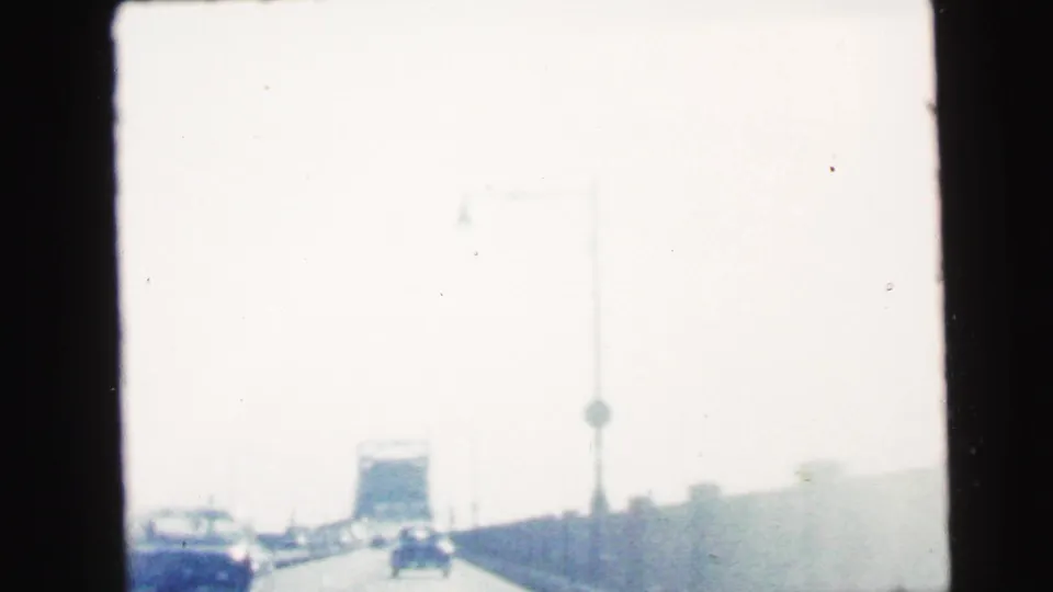 1964 home-movie frame from Camden, New Jersey showing a bright highway view from inside a moving car, with traffic and roadside structures ahead.