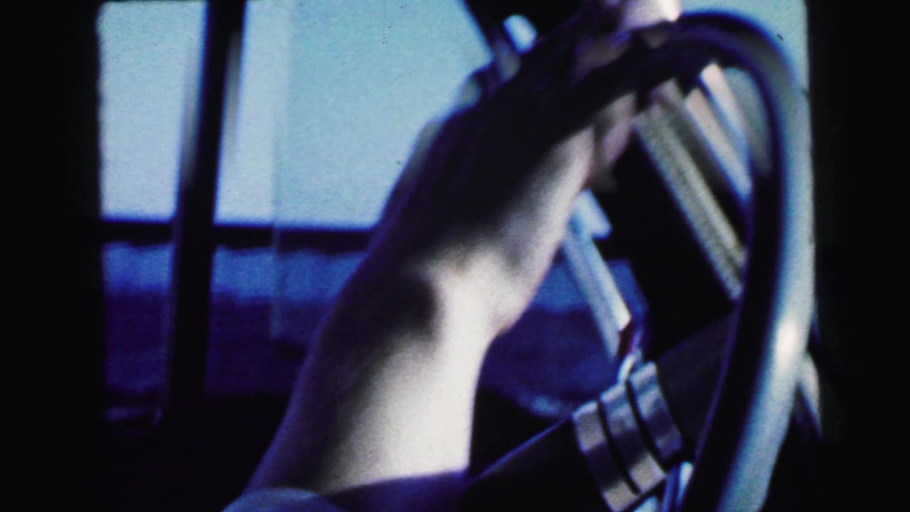 Close 1964 car-interior view showing the driver's hand on the steering wheel beside a blue-tinted side window.