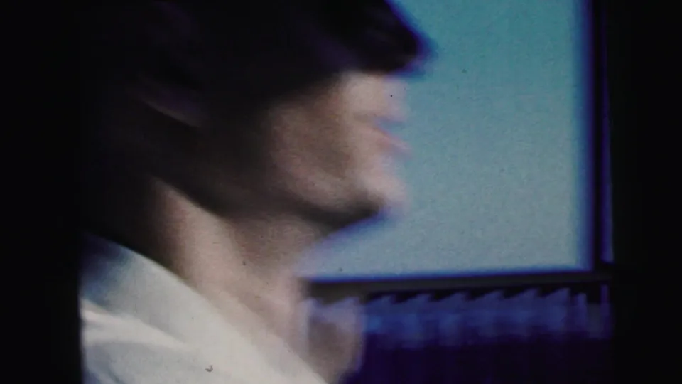 Soft-focus 1964 home-movie frame of a smiling man in sunglasses seated in the car, filmed from the passenger side.