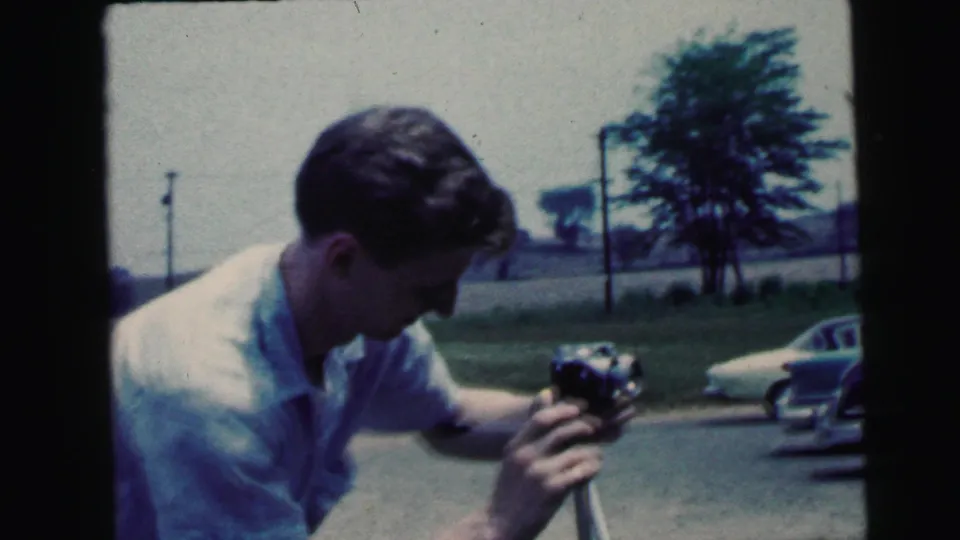 1964 Camden home-movie frame of a young man in a white shirt adjusting a tripod-mounted camera near parked cars and grass.