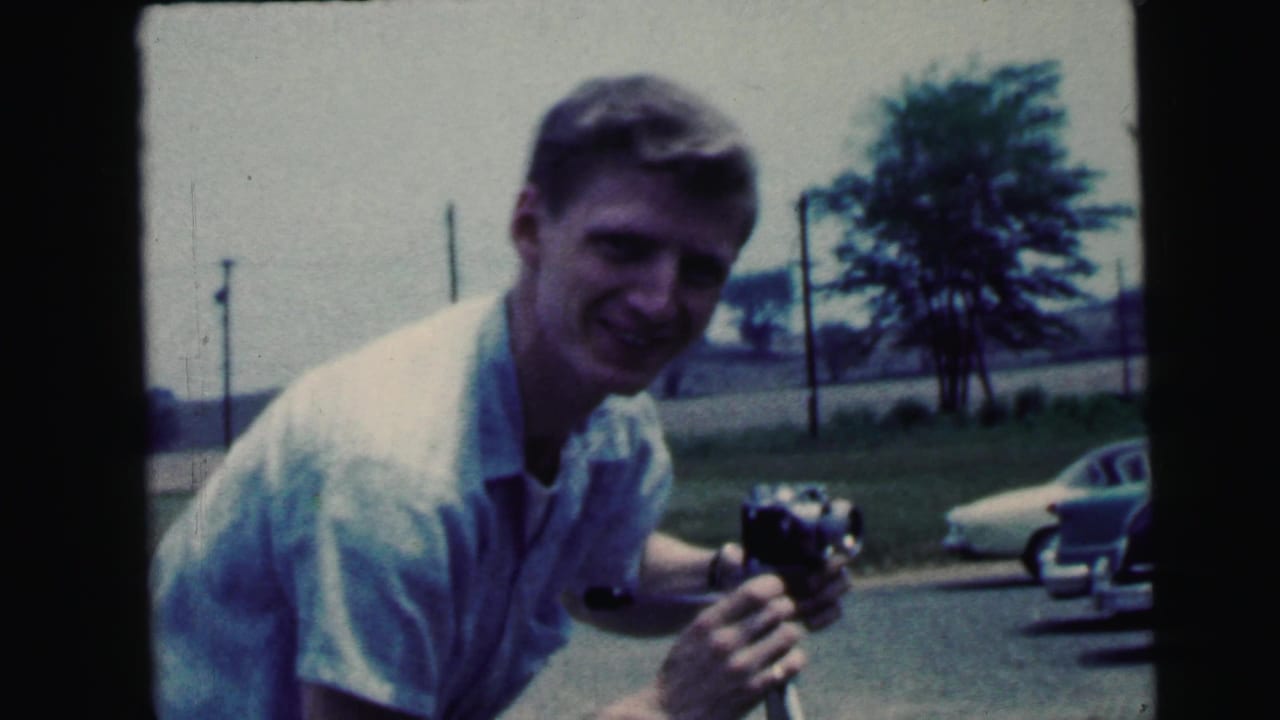 Young man smiling outdoors while holding a small camera near parked cars.