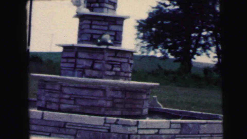 Clearer 8mm frame of a tiered stone monument or fountain structure in Camden, with grass, trees, and paved edging visible.