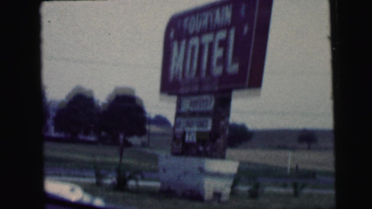 Tall Fountain Motel roadside sign seen from a moving car beside open ground and trees.