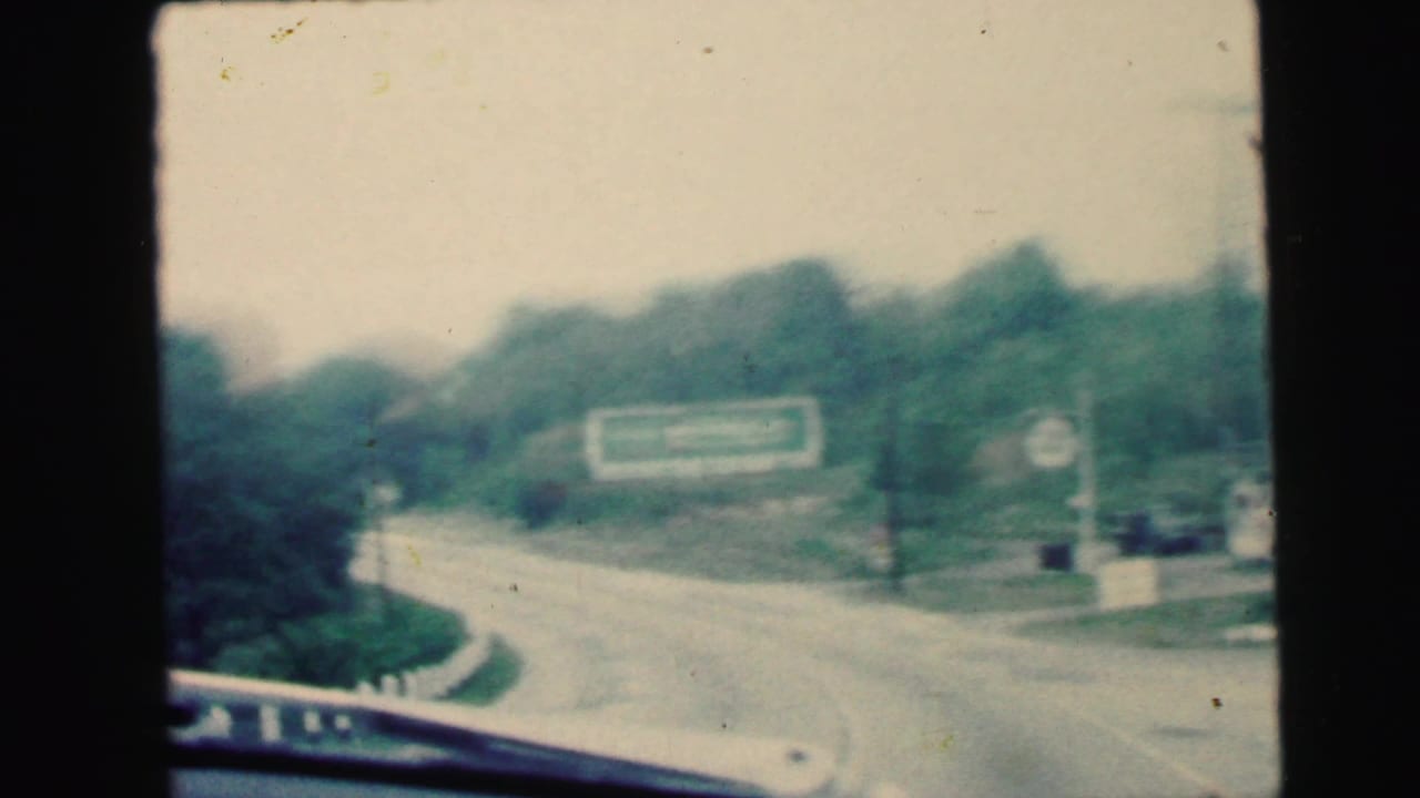 Moving-car view in the Camden, New Jersey clip, with a curving road, windshield edge, trees, and a green roadside billboard ahead.