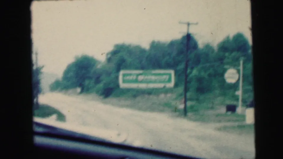 1964 8mm road-trip frame with a green billboard, trees, utility pole, roadside verge, and car-window edge in Camden, New Jersey footage.