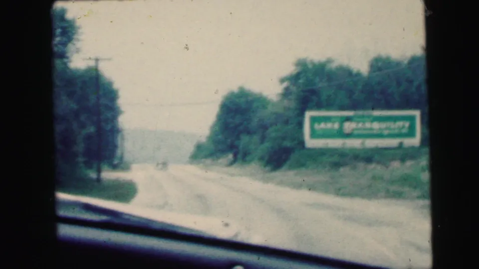 Final frame of the Camden, New Jersey road-trip clip, showing a green billboard, trees, road, and windshield edge in soft 8mm color.