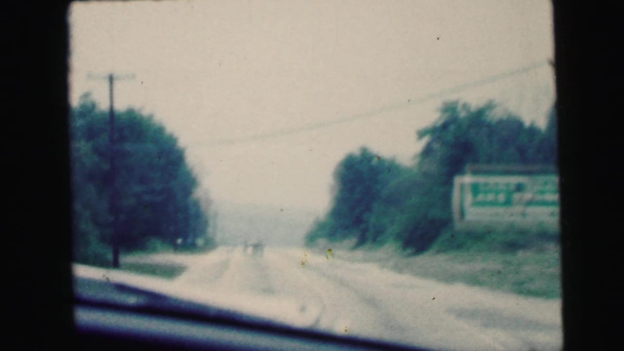 1964 Camden, New Jersey road-view footage showing a two-lane highway, trees, windshield edge, and a green roadside sign.