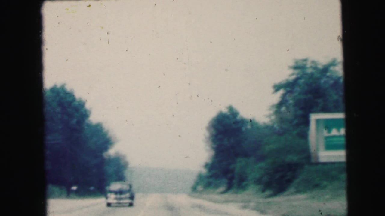 1964 Camden, New Jersey windshield POV with an approaching car, roadside trees, pale sky, and a partly cropped sign.