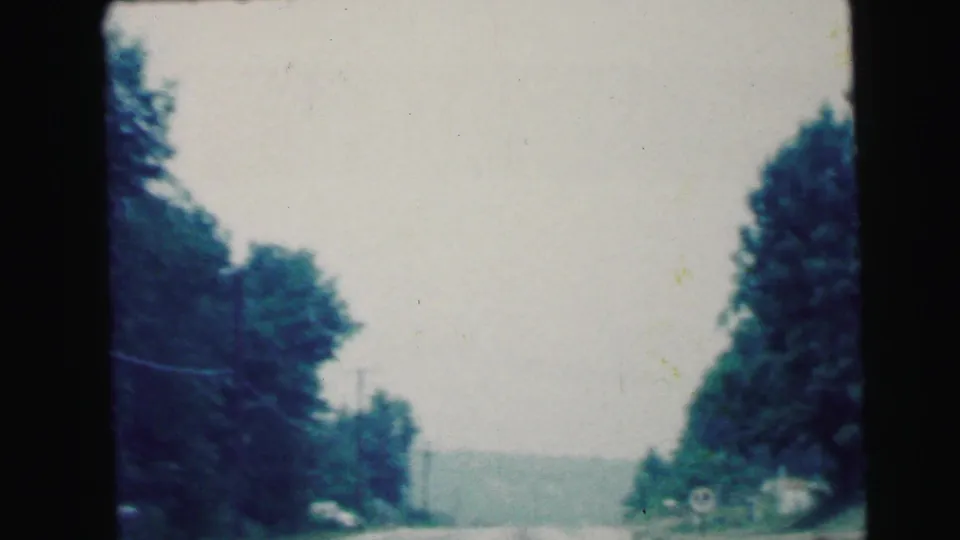 1964 Camden, New Jersey windshield view of a two-lane road bordered by trees, distant hills, and small roadside signs.