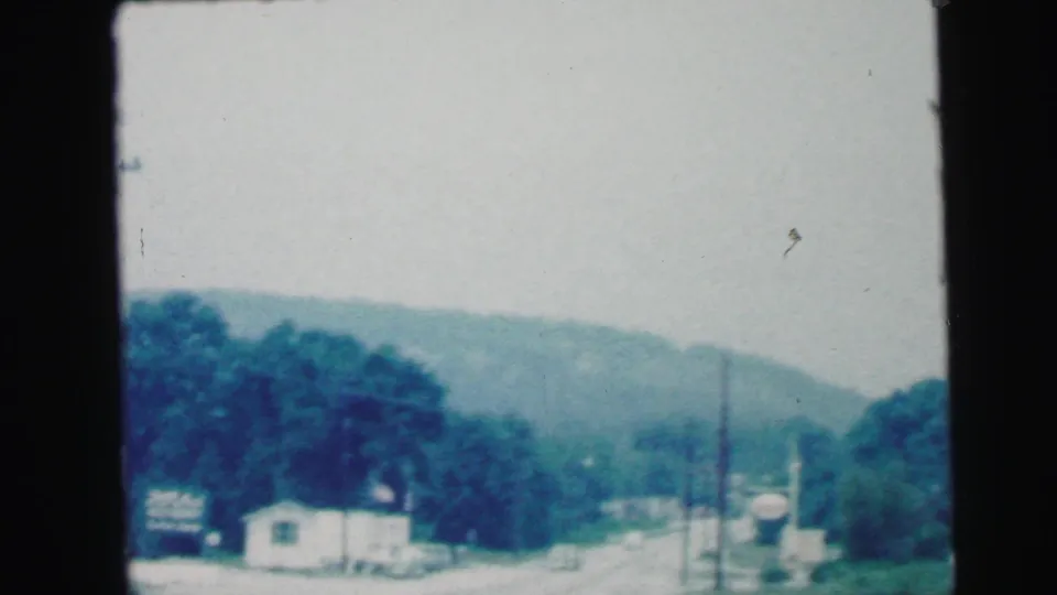 1964 8mm driving view in Camden, New Jersey, with a curving road, small roadside building, utility poles and wooded hills.