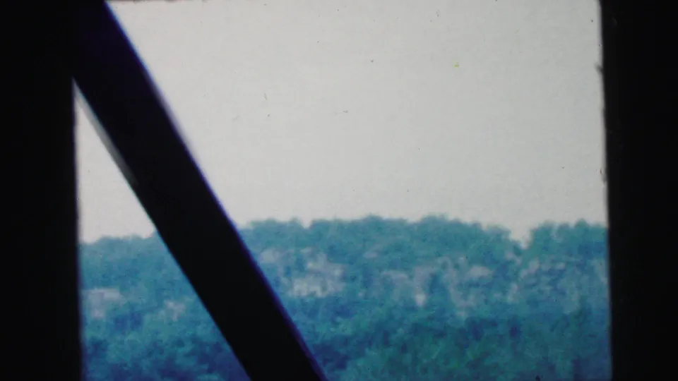 End frame from 1964 Camden 8mm footage, with a dark windshield pillar over wooded hills, roadside greenery and an overcast sky.