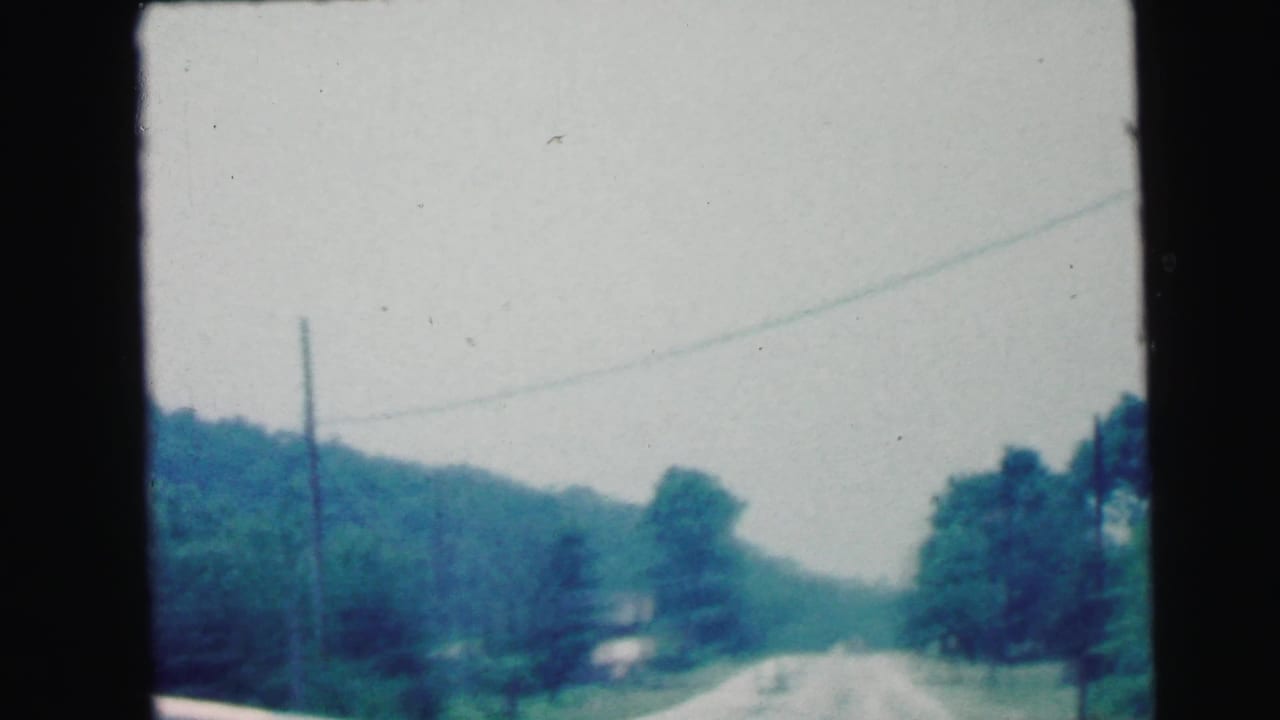 1964 rural New Jersey driving view from a moving vehicle, with a road, utility poles, trees, and wooded hills ahead.