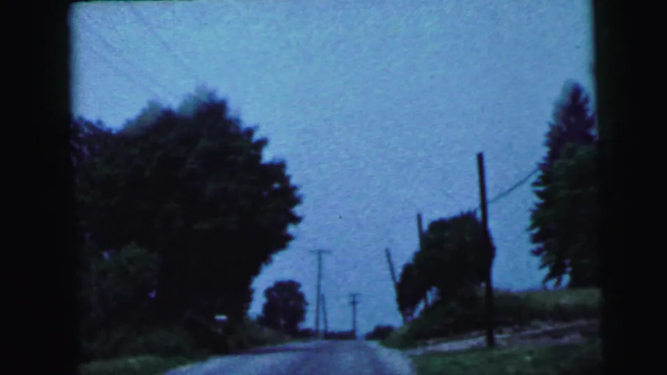 Curved rural two-lane road, deciduous tree line, and utility poles with wires, shot from inside a moving car, 8mm circa 1964 New Jersey.