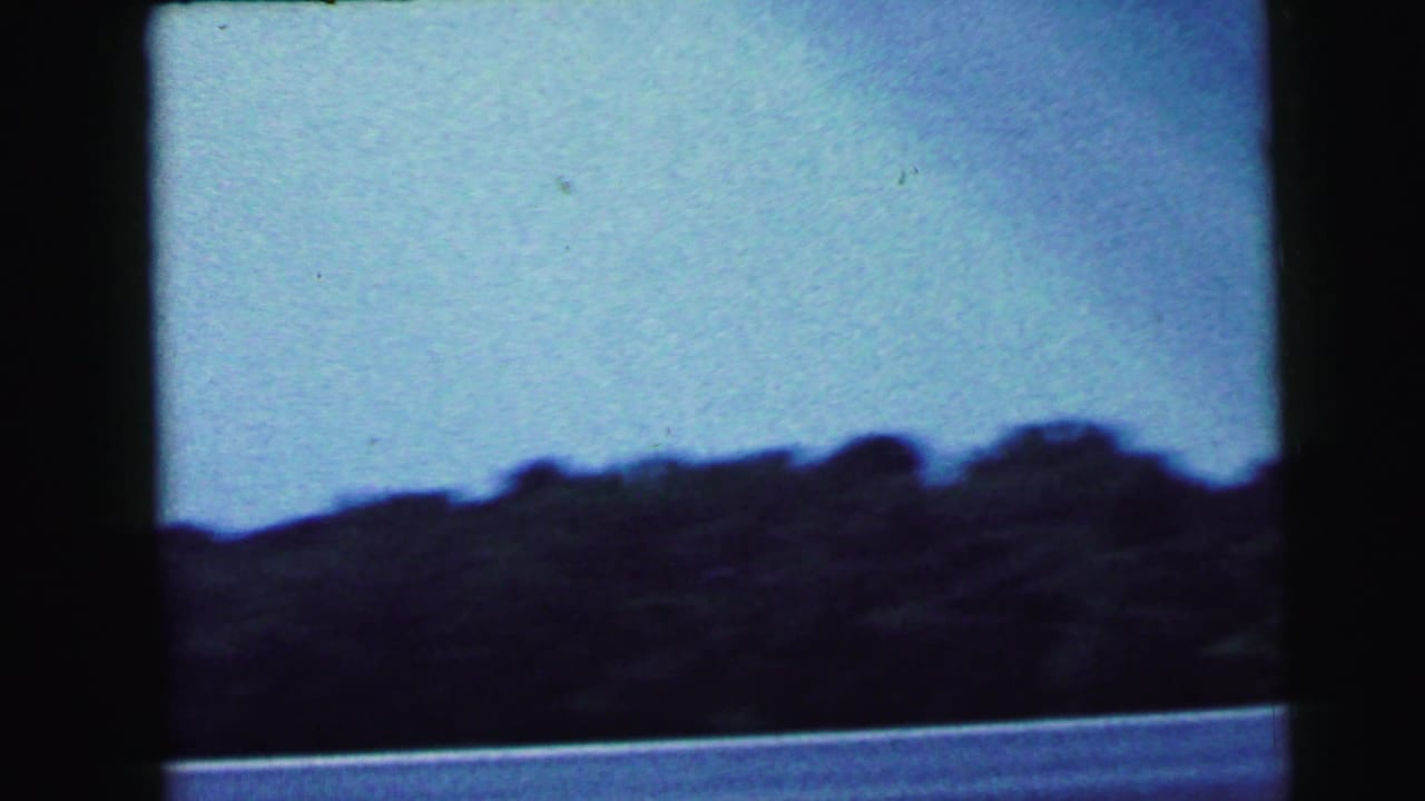 8mm home movie still from 1964 showing a silhouetted treeline against a pale blue sky, likely filmed from a moving vehicle in New Jersey.