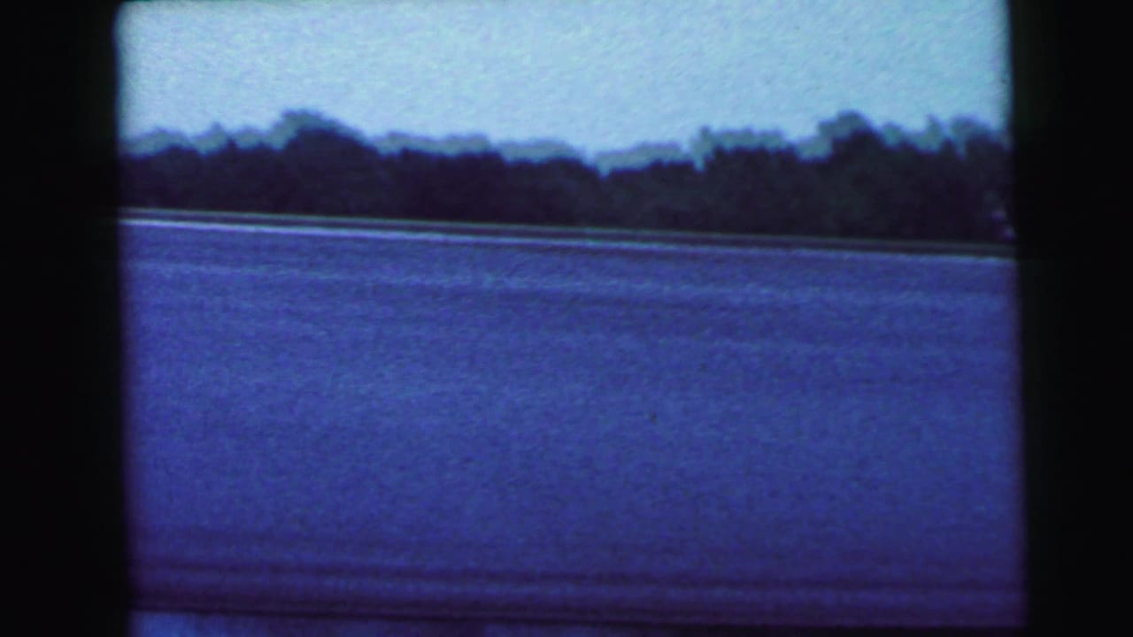 Calm body of water beneath a blue sky with a dark treeline silhouette at the horizon, 8mm film still with strong blue color shift.