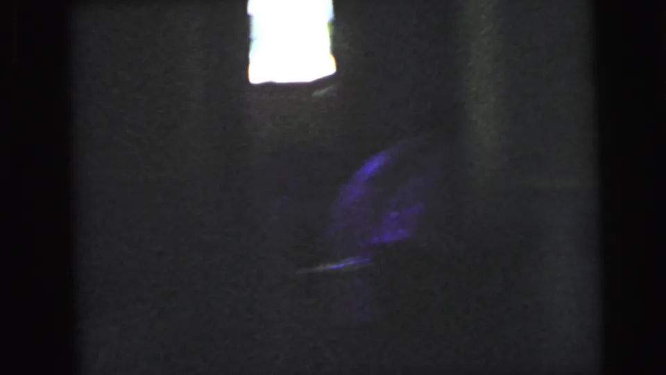 Dark interior space with an overexposed window highlight and a shadowed mass that may indicate a partially visible person, 8mm film circa 1964.
