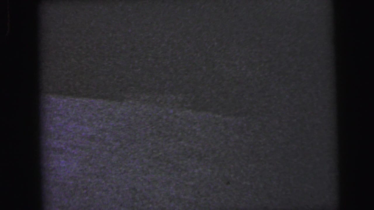 Nearly black 8mm film frame with faint horizontal surface texture in heavy shadow; interior setting almost entirely obscured by underexposure.