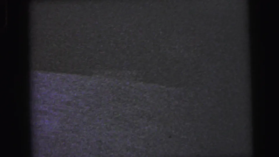 Severely underexposed opening frame from a 1964 8mm home movie; a horizontal interior surface is barely visible through dense grain and near-total darkness.