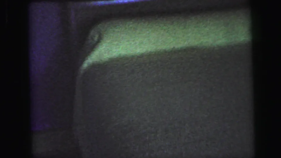 8mm home movie frame from 1964 showing a curved upholstered surface lit by green and purple light, possibly a pillow or mattress edge in a darkened bedroom.