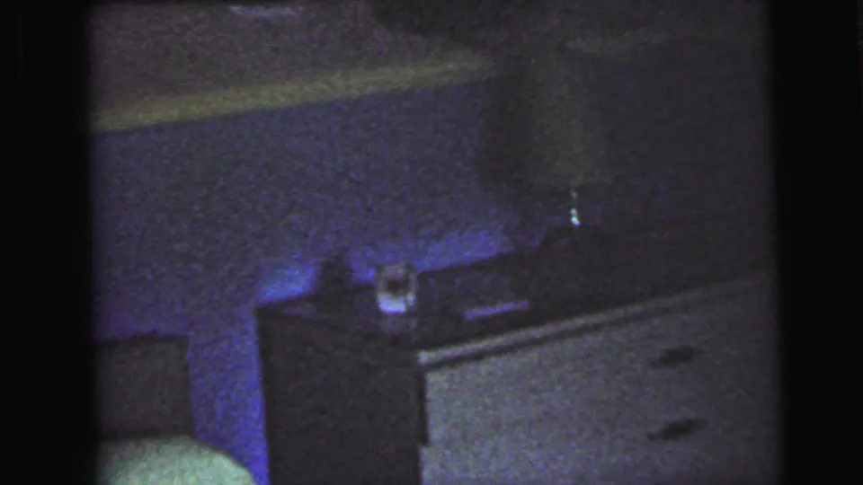 1964 8mm home movie bedroom frame showing a low chest of drawers with a small object on top, blue-purple light in the lower corner, and the edge of a bed or headboard in warm tones above.