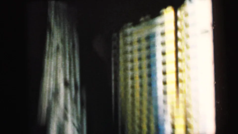 1964 8mm home movie frame capturing sheer fabric alongside a yellow patterned textile — possibly a curtain and bedspread — in a brighter portion of the bedroom interior sequence.