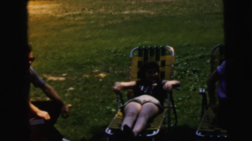 A woman relaxes in a webbed folding lawn chair on a backyard lawn, with partial figures at frame edges; consistent with mid-1960s suburban domestic footage from Camden, New Jersey.
