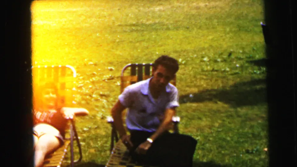 A man in a white short-sleeved shirt sits in a folding lawn chair on a backyard lawn with lens flare at frame left; consistent with 1964 8mm home movie footage from Camden, New Jersey.