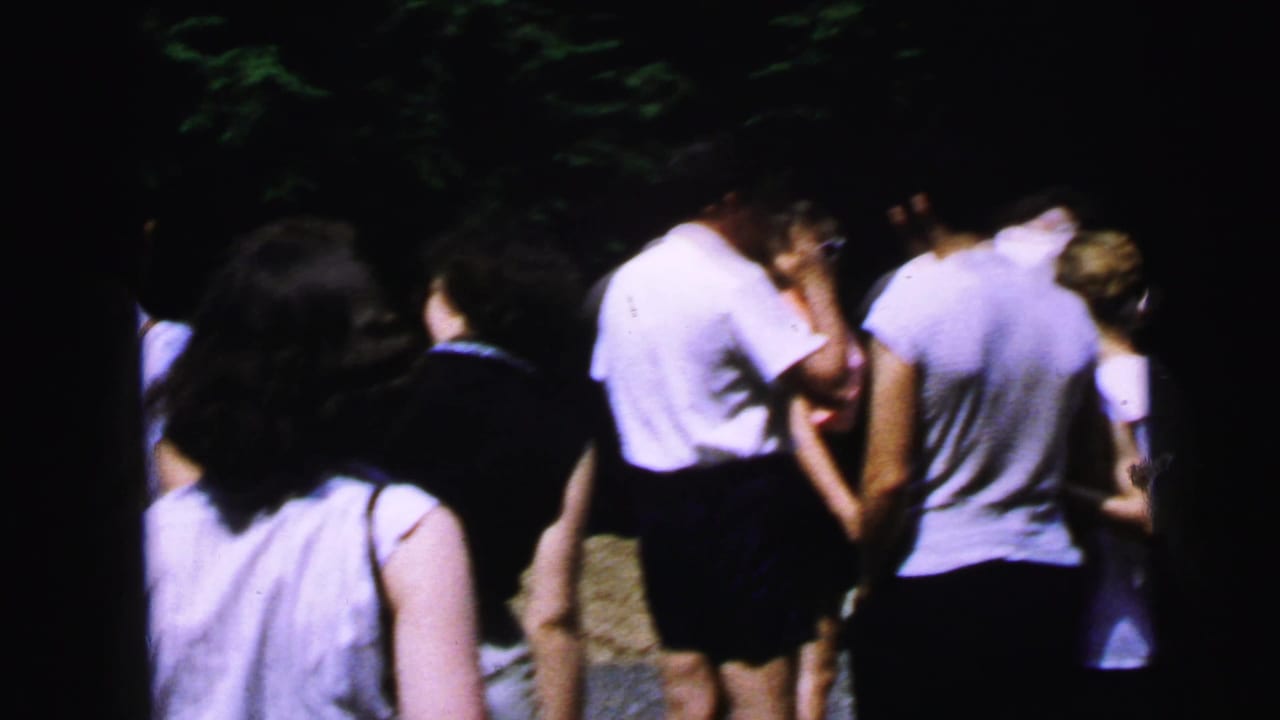 Group of adults seen mostly from behind, wearing white shirts and dark shorts, gathered outdoors near trees.