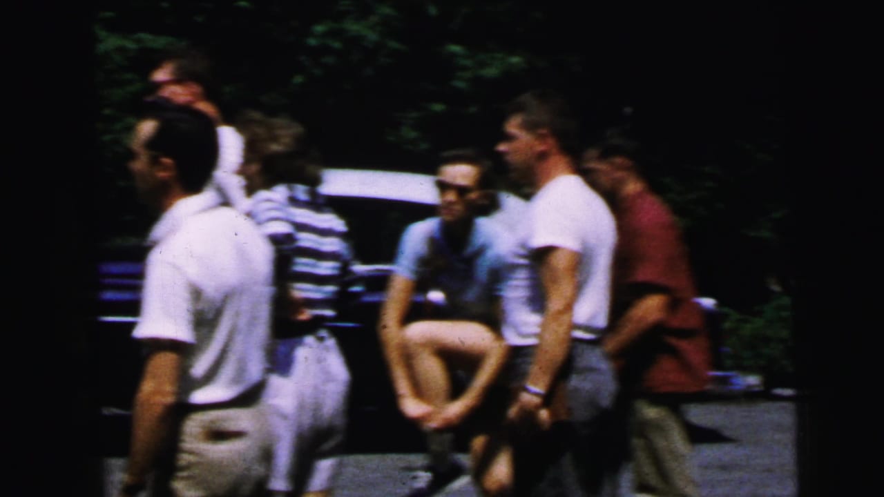 Group of young adults in casual summer clothing gathered outdoors in a wooded area near a parked dark vehicle.