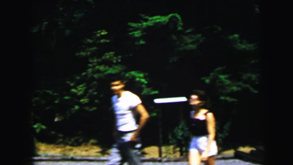 A man in a white t-shirt and a woman walk across a sun-lit outdoor area; the top of a yellow novelty vehicle is partially visible below, wooded setting, circa 1964.