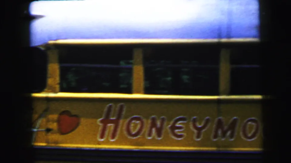 Close view of a yellow novelty vehicle's side panel displaying 'HONEYMO[ON]' in decorative script with a red heart emblem; two young adults visible above against green trees, circa 1964.